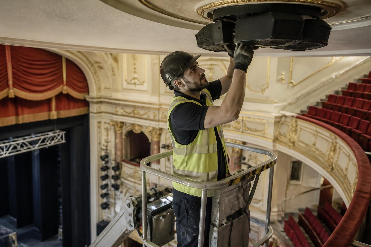 Sound installer on a mobile platform fitting a ceiling speaker cluster above the stalls of a refurbished UK theatre
