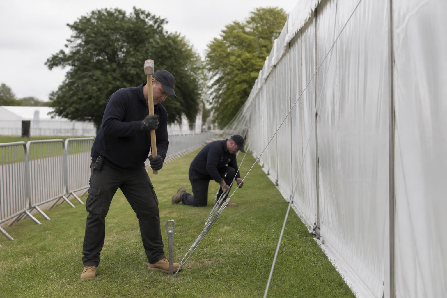 Two crew members driving ground stakes and tensioning guy lines beside a clearspan structure at a UK county show