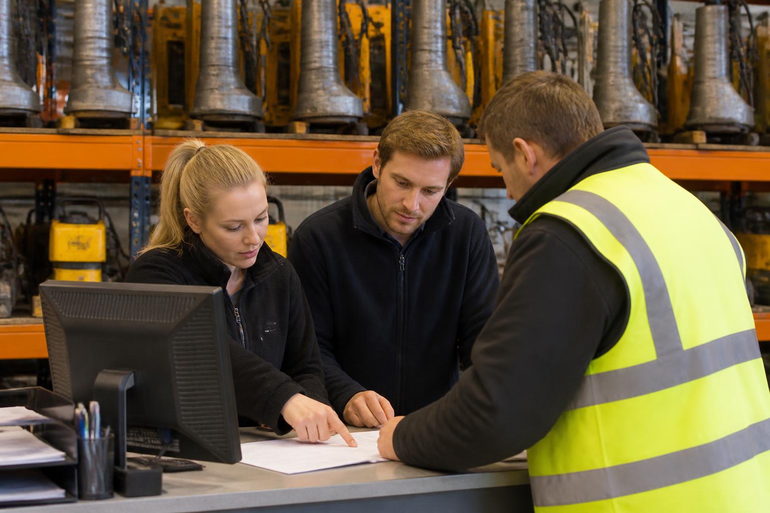 Hire desk staff going through paperwork with a site manager at a busy UK plant and tool hire counter, with breakers and compactors racked behind them