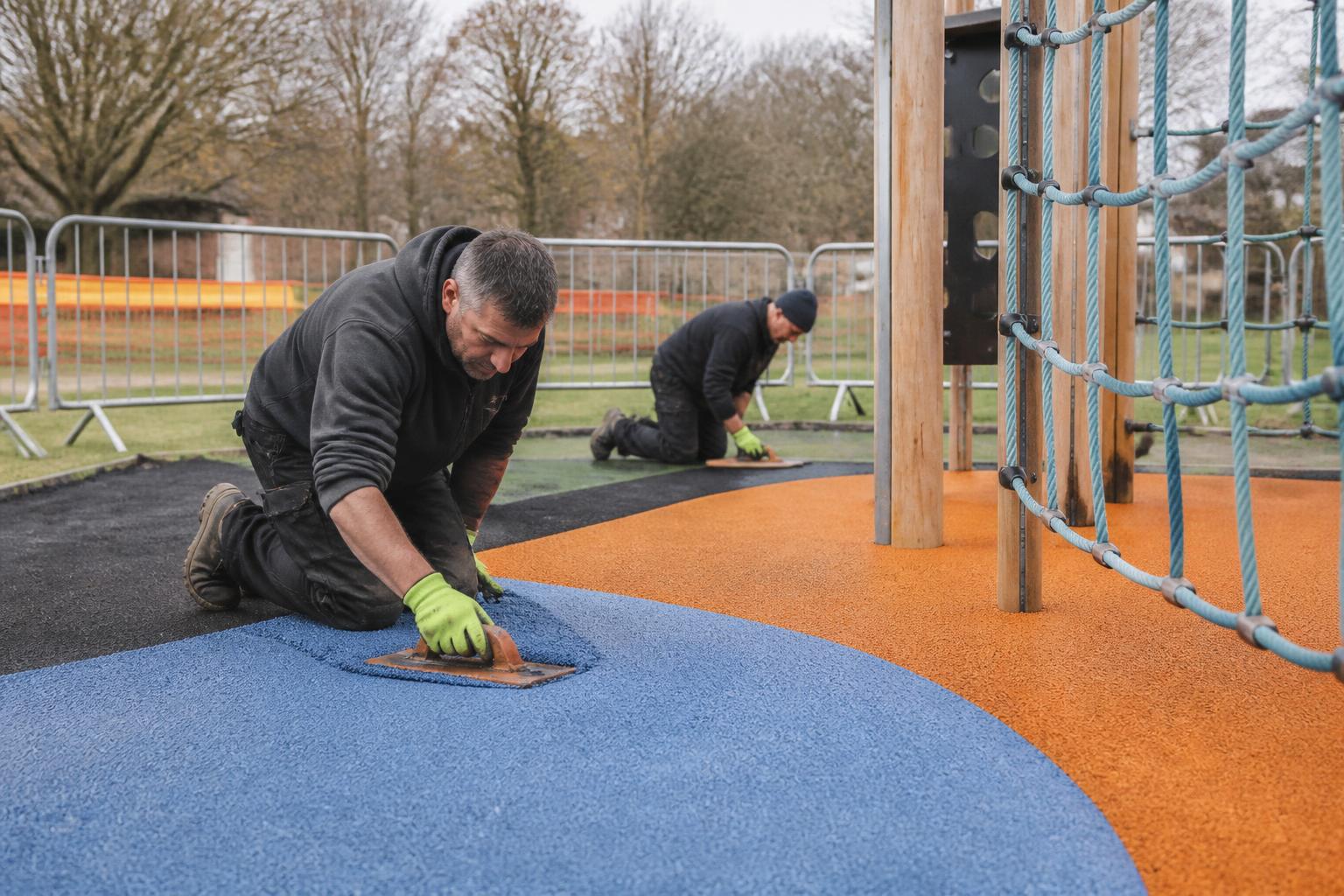 Two installers laying colourful wet pour safety surfacing around new climbing equipment in a UK park playground, temporary barriers set around the site